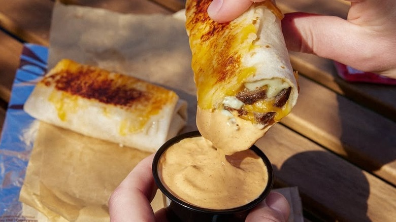 A close-up of a hand dipping one of Taco Bell's cheesy dipping burritos into a pot of sauce, with another burrito on the table in the background